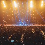 Esta imagem mostra uma apresentação ao vivo em um grande palco, iluminado por luzes amarelas e azuis. A banda está performando no centro, com o vocalista destacando-se no meio. O público, numeroso e entusiasmado, está de pé e com as mãos levantadas, claramente envolvido e aproveitando o espetáculo. Luzes de palco adicionais e efeitos visuais acrescentam um clima energético ao ambiente, enquanto mesas à frente da plateia sugerem um formato de show que combina áreas de assentos com áreas de pé.