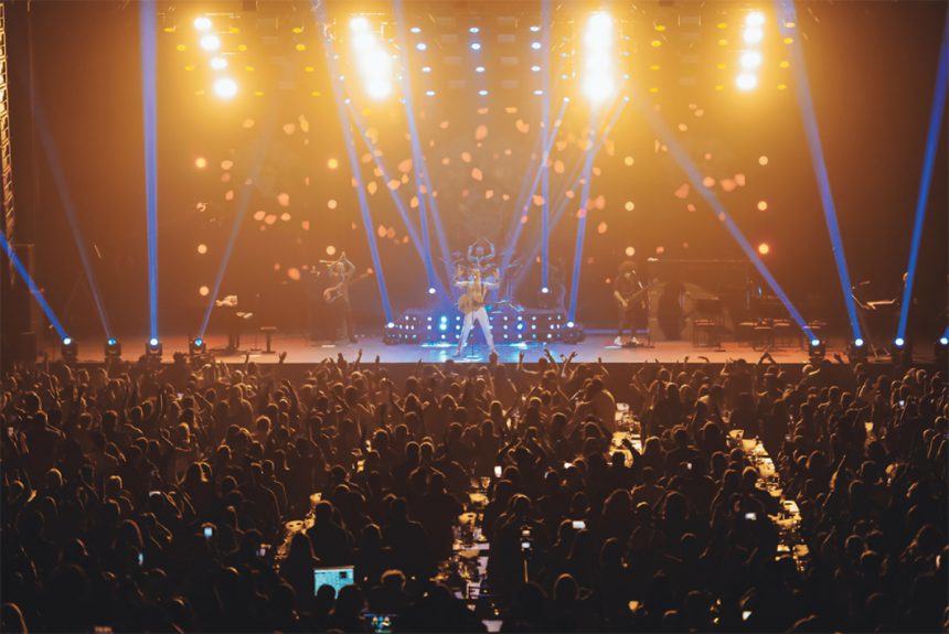 Esta imagem mostra uma apresentação ao vivo em um grande palco, iluminado por luzes amarelas e azuis. A banda está performando no centro, com o vocalista destacando-se no meio. O público, numeroso e entusiasmado, está de pé e com as mãos levantadas, claramente envolvido e aproveitando o espetáculo. Luzes de palco adicionais e efeitos visuais acrescentam um clima energético ao ambiente, enquanto mesas à frente da plateia sugerem um formato de show que combina áreas de assentos com áreas de pé.