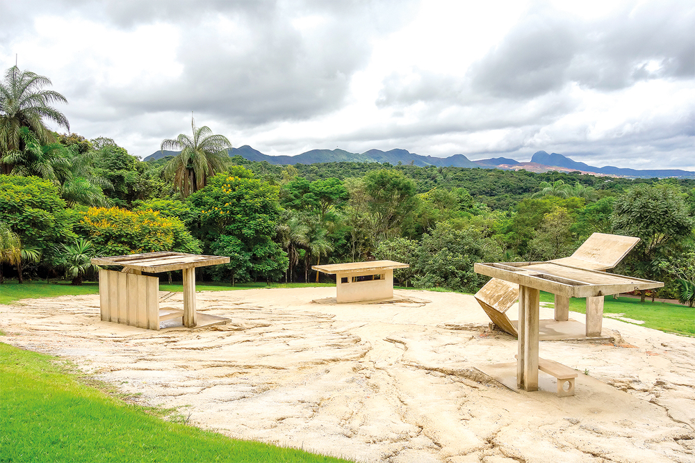 Escultura ao ar livre no Instituto Inhotim, em Brumadinho, com mesas e cadeiras de concreto em meio a uma paisagem exuberante de vegetação e montanhas ao fundo.