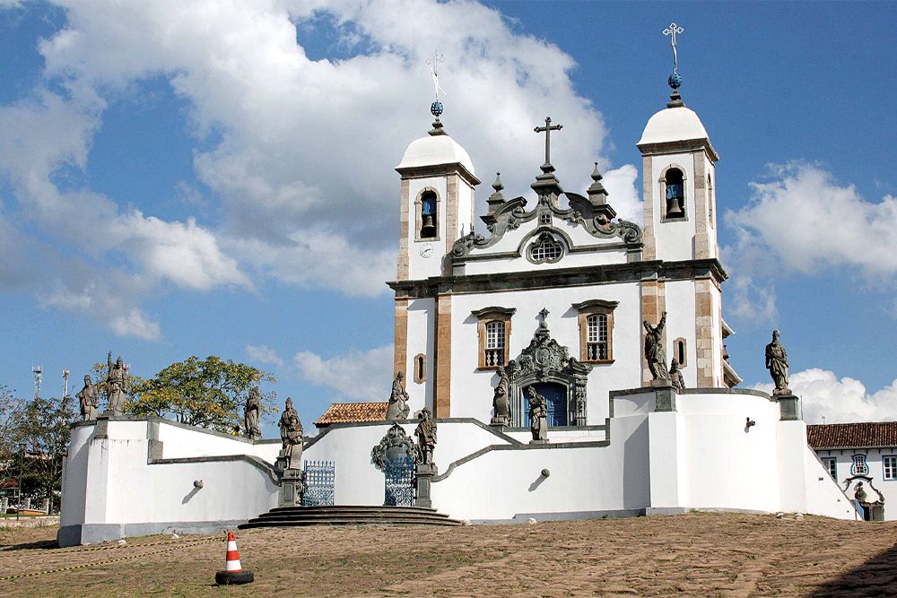 Fachada do Santuário do Bom Jesus de Matosinhos, em Congonhas, Minas Gerais, com esculturas de profetas de Aleijadinho em primeiro plano, sob um céu azul com nuvens.