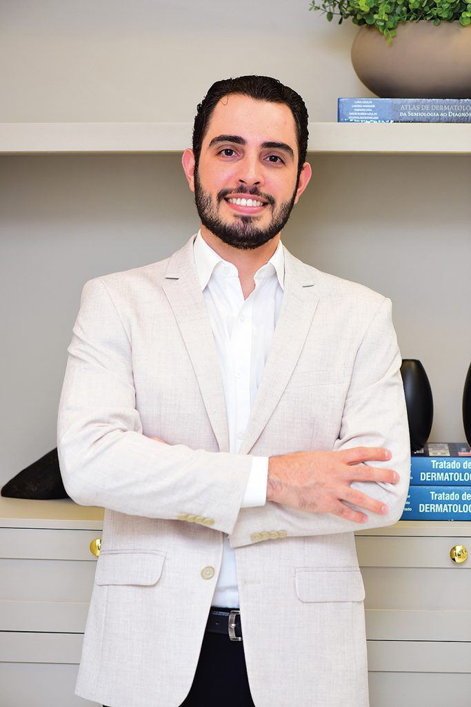 Homem sorridente de terno bege claro, posando diante de uma prateleira com livros de dermatologia e decoração.